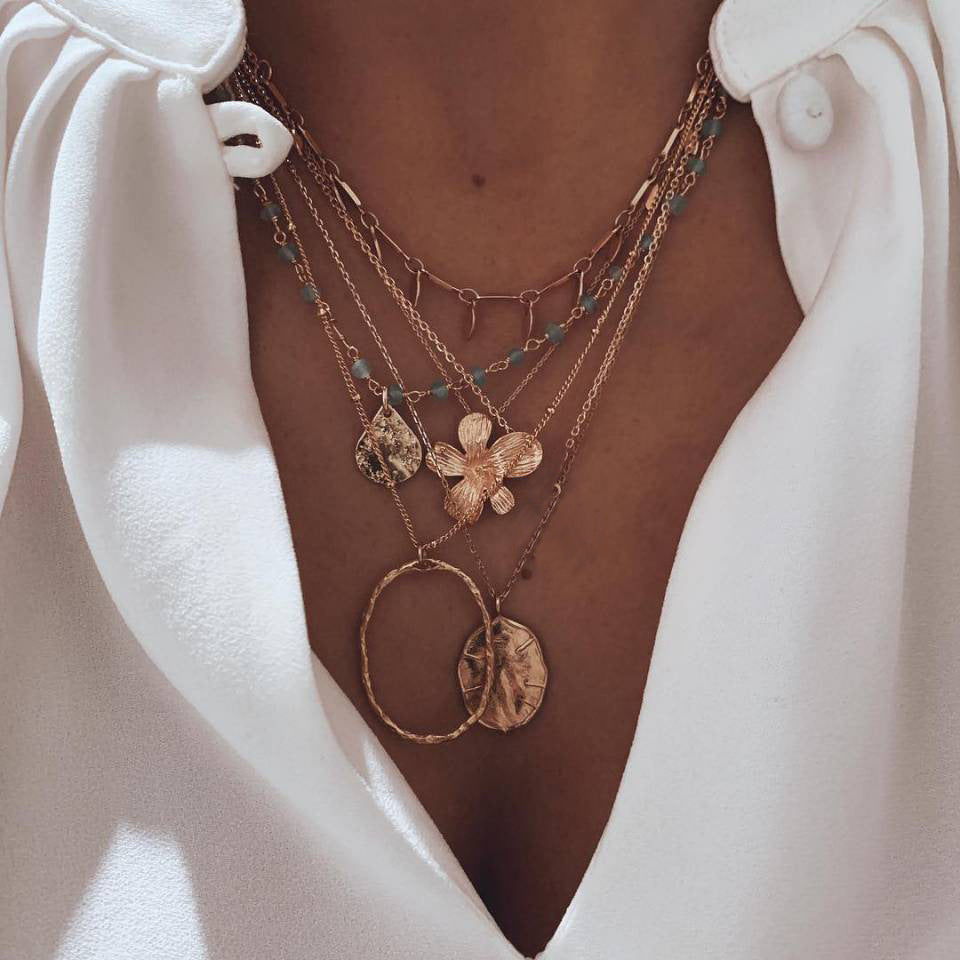 Layered gold-tone necklaces with flower, leaf and circular pendants, worn with a white shirt